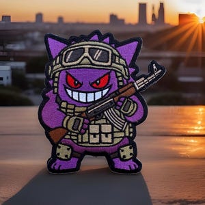 May include: A purple embroidered patch featuring a cartoon creature with red eyes and a wide grin. The character wears a tan tactical vest, knee pads, and goggles on its head while holding a brown and black rifle across its chest.