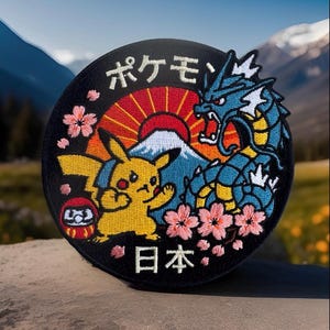 May include: Round embroidered patch featuring Pikachu and a blue dragon with Japanese text. The design includes a red sun, a mountain, and pink cherry blossoms on a black background. The patch is likely for clothing or accessories.