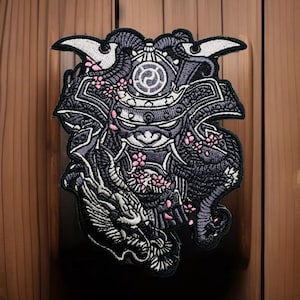May include: A black and white embroidered patch featuring a samurai warrior with a dragon and cherry blossoms. The samurai is wearing a helmet with a circular design on the front. The patch is surrounded by a white border.