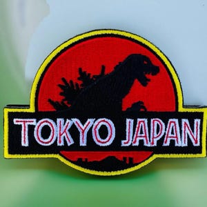 May include: A black and yellow embroidered patch with a silhouette of Godzilla in front of a red sun. The text "TOKYO JAPAN" is written in white with a red outline on a black background.