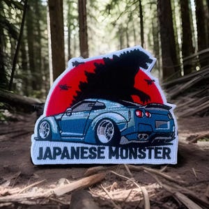 May include: A white embroidered patch with a blue car, a red sun, and a black silhouette of Godzilla. The text "JAPANESE MONSTER" is printed below the car.