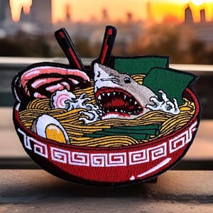 May include: A red and white embroidered patch of a shark swimming in a bowl of ramen noodles. The shark has a large open mouth with sharp teeth. The ramen noodles are yellow and the bowl has a red rim with a white design.