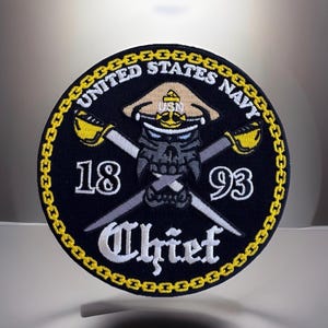 May include: A round, navy blue patch with a gold chain border. The patch features a skull wearing a hat, crossed swords, and the text "UNITED STATES NAVY", "18", "93", and "Chief".