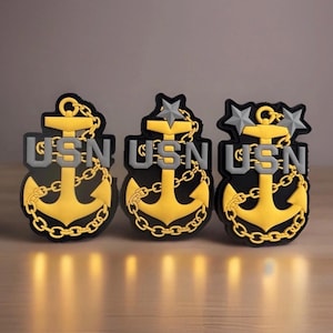 May include: Three black and gold anchor emblems with the letters "USN" in gray. The anchors have gold chains and are topped with stars. The emblems are on a wooden surface.