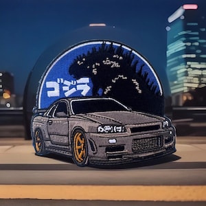 May include: A gray embroidered patch featuring a black silhouette of Godzilla standing over a gray Nissan Skyline GT-R. The patch has a blue background with white text that reads "ゴジラ" in Japanese.