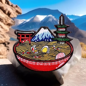 May include: A red bowl of ramen noodles with shrimp, a boiled egg, and a swirl of pink fish cake. The bowl is decorated with a mountain, a pagoda, and a torii gate.