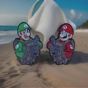 May include: Two embroidered patches featuring cartoon characters Mario and Luigi in military gear, holding rifles. Mario wears a red hat and shirt, while Luigi sports a green hat and shirt. Both patches are set against a beach backdrop with sand and ocean.