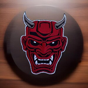 May include: Red embroidered iron-on patch of a Japanese demon mask with sharp teeth and horns.