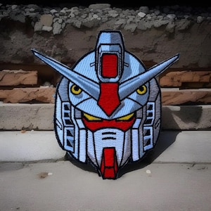 May include: A close-up of a blue and red embroidered patch featuring the head of a robot with a large visor and yellow eyes. The patch is on a gray concrete surface.
