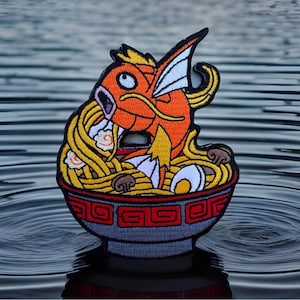 May include: Embroidered patch featuring an orange cartoon fish with yellow fins and whiskers, eating ramen noodles from a gray bowl with a red rim. The patch is set against a water background.