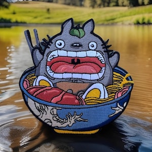 May include: Embroidered patch featuring a gray cartoon character with a wide open mouth, sitting in a blue bowl of ramen. The ramen includes noodles, meat, and eggs. Chopsticks are placed on the side of the bowl. The patch is set against a blurred background of water and greenery.