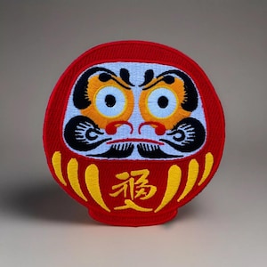 May include: A red and white embroidered daruma doll with a black and yellow face. The doll has a yellow "福" character on the front.