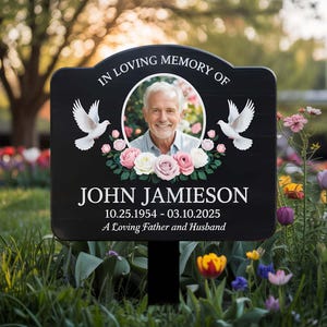 May include: Black memorial sign with a white border and rounded top. The sign features a photo of a man in a white shirt, surrounded by pink and white roses and two white doves. The text reads "In Loving Memory Of John Jamieson 10.25.1954 - 03.10.2025 A Loving Father and Husband".