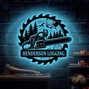 Op de afbeelding: Een rond metalen wandbord met blauwe led-achtergrondverlichting met een kettingzaag, dennenbomen, bergen en boomstamdoorsneden. De tekst HENDERSON LOGGING staat gecentreerd in een horizontale balk onderaan het zaagbladontwerp.