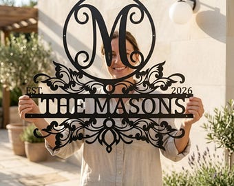 May include: Black metal family name sign with the name "THE MASONS" and the year "2026". The sign features a large letter "M" inside a circle, with decorative scrollwork. The sign is held up by a person.