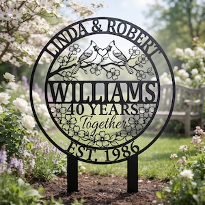 May include: A circular black metal garden sign featuring the text LINDA & ROBERT, WILLIAMS, 40 YEARS Together, and EST. 1986. The design includes two birds perched on a branch with a heart between them, surrounded by floral cutouts and two stakes.