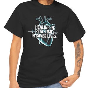 May include: Black t-shirt featuring a stylized, glowing blue anatomical heart graphic. The text "HEALING IN REAL-TIME IR SAVES LIVES" is printed above the heart, with a heartbeat line. The shirt is a crew neck.