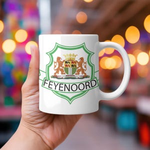 Puede incluir: Taza de cerámica blanca con el emblema del Feyenoord, un escudo verde y blanco con dos leones, una corona y la palabra "FEYENOORD". La taza tiene un asa curva y está sujeta con una mano.