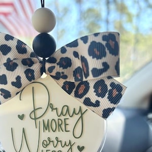 May include: A car charm with a leopard print bow and a clear acrylic disc. The disc has the words "Pray More Worry Less" in green script. The charm is suspended by a black string with two beads.