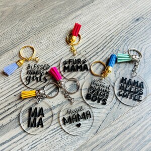 May include: A collection of clear acrylic keychains with gold key rings and colorful tassels. Each keychain has a different black text design, including phrases like "Blessed with Girls" and "Mama Needs Coffee."