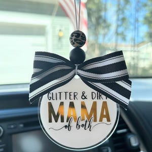 May include: A round car air freshener with the text "Glitter & Dirt MAMA of both" in gold and black. It has a black and white striped bow and a leopard print bead.