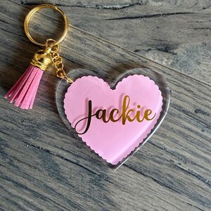 May include: A heart-shaped keychain with a pink center and the name "Jackie" in gold script. The keychain has a gold-colored ring, chain, and a pink tassel. The background is a wooden surface.