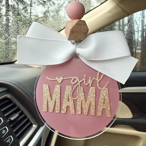 May include: A circular car air freshener ornament featuring a dusty rose background with the text girl MAMA in gold glitter script and block lettering. The ornament is topped with a white ribbon bow, a wooden bead, and a dusty rose silicone bead.