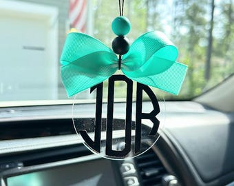 May include: A circular clear acrylic car ornament featuring a black monogram with the letters JDB. The ornament is topped with a bright turquoise grosgrain ribbon bow, a turquoise silicone bead, and a smaller black bead, all hanging from a black cord.