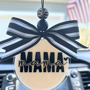May include: A car charm with a cream-colored circular pendant featuring the word "MAMA" in black, with a small heart. It is adorned with a black and white striped bow and two beads. The charm hangs from a black string.