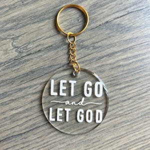May include: Clear acrylic keychain with gold-colored hardware. The keychain has a round pendant with the words "LET GO and LET GOD" in white lettering. The keychain is on a wooden surface.