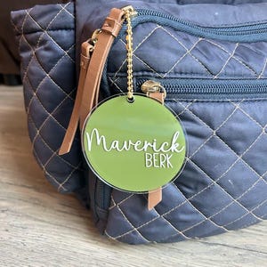 May include: A circular olive green acrylic bag tag featuring the white text Maverick BERK, attached to a navy blue quilted backpack with a gold-toned metal ball chain. The tag has a smooth, glossy finish and is suspended from a zipper pull.