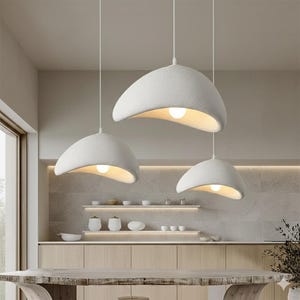 May include: Three white, textured pendant lights hang from the ceiling. Each light has a curved, organic shape with a warm, glowing bulb inside. The lights are suspended above a wooden table in a modern kitchen setting.