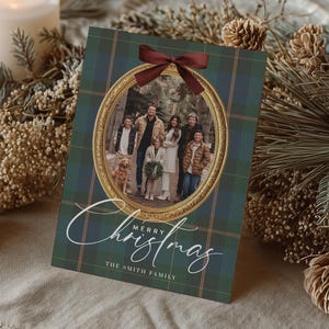 May include: A Christmas card with a family photo inside a gold frame, set against a green and blue plaid background. The card is adorned with a burgundy bow and the words "Merry Christmas" in elegant script. Two lit candles and festive greenery surround the card.