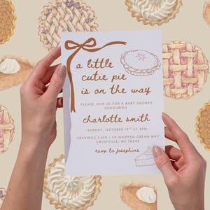 May include: A baby shower invitation held by hands, featuring the text "a little cutie pie is on the way." The invitation has a brown ribbon design and a pie illustration. The background has watercolor pie illustrations.