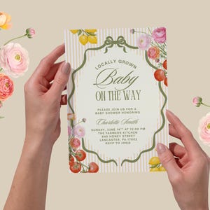 May include: A baby shower invitation held by hands, featuring the text "Locally Grown Baby On The Way." The invitation has a decorative border with flowers, lemons, and tomatoes. The background is a light beige with vertical stripes.