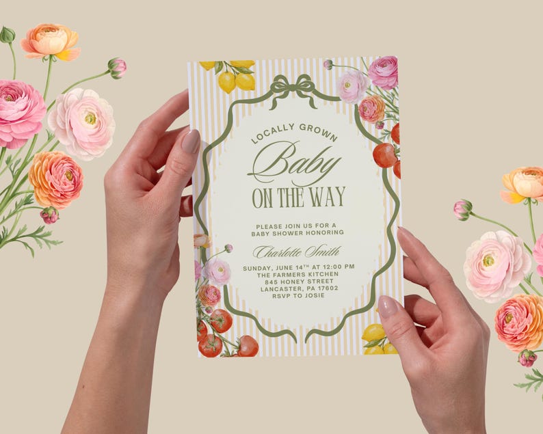 May include: A baby shower invitation held by hands, featuring the text "Locally Grown Baby On The Way." The invitation has a decorative border with flowers, lemons, and tomatoes. The background is a light beige with vertical stripes.