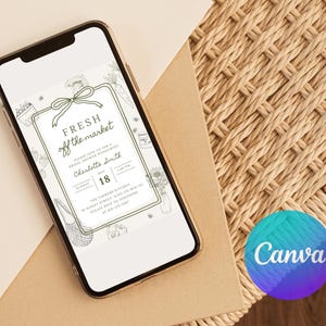 May include: A smartphone displays a digital invitation with the text "FRESH off the market." The invitation features a green border with illustrations of vegetables and flowers. The phone rests on a light brown surface next to a woven mat.