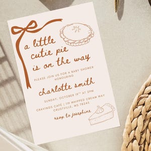 May include: A baby shower invitation with a light beige background and brown text and illustrations. The text reads "a little cutie pie is on the way" and includes details for the event honoring Charlotte Smith.