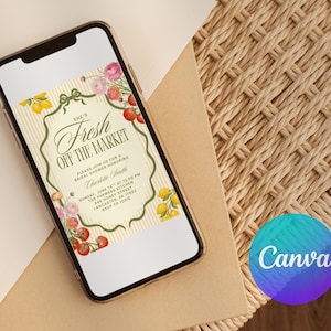 May include: A smartphone displays a bridal shower invitation with the text "She's Fresh Off The Market." The invitation features floral and fruit illustrations in pink, red, and yellow, set against a cream background with vertical stripes. The phone rests on a woven surface.