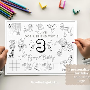 Op de afbeelding: Een zwart-wit gepersonaliseerde verjaardagskleurplaat met stripfiguren en de tekst "YOU'VE GOT A FRIEND WHO'S" en "Ryan's 3rd Birthday". Een kind kleurt de pagina met een paars potlood. Kleurpotloden liggen verspreid.