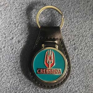 May include: Black leather keyring with a silver ring and a teal and red emblem that reads "CRESSIDA". The emblem has a silver border and a stylized design.