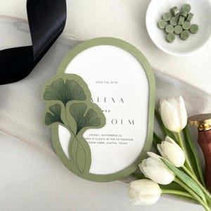 May include: An olive green wedding invitation with a ginkgo leaf design. The invitation has the text "SAVE THE DATE" and the names "ALEXA AND COLM" along with the date and location. White tulips and green wax seals are also in the image.