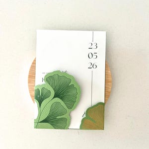 Peut inclure: Une carte rectangulaire blanche affichant la date 23 05 26 en noir. La carte est ornée d'illustrations de feuilles de ginkgo vertes dans les coins inférieurs gauche et droit, avec une surface texturée et une ligne noire verticale.