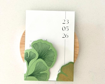 May include: A white rectangular card featuring the date 23 05 26 in black text. The card is decorated with green ginkgo leaf illustrations on the bottom left and right corners, with a textured surface and a vertical black line near the date.