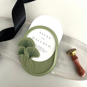Könnte beinhalten: Eine elegante Hochzeitseinladung mit einem grünen Blattdesign und den Namen "ALEXA und MALCOLM" auf einer weißen ovalen Karte. Die Einladung ist über einem grünen ovalen Rahmen angeordnet. Ein goldener Wachssiegel und ein schwarzes Band sind ebenfalls sichtbar.