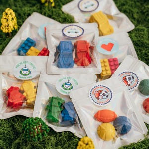 May include: Multiple clear plastic pouches containing assorted shaped wax crayons. The pouches feature circular stickers with various designs, including a robot with the text 'YOU'RE BOT-TASTIC THANK YOU' and a sports theme with 'YOU'RE AN ALL-STAR THANK YOU'.