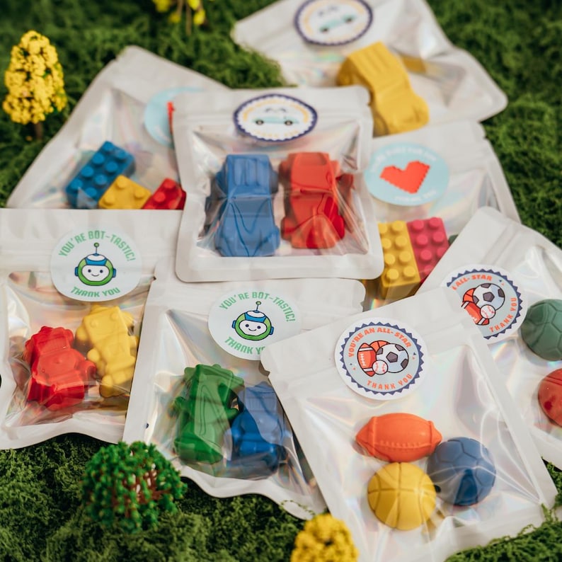 May include: Multiple clear plastic pouches containing assorted shaped wax crayons. The pouches feature circular stickers with various designs, including a robot with the text 'YOU'RE BOT-TASTIC THANK YOU' and a sports theme with 'YOU'RE AN ALL-STAR THANK YOU'.