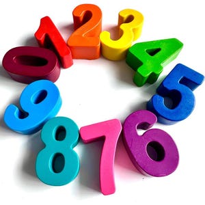 May include: A set of ten individual numbers from zero to nine, each crafted in a distinct solid color including red, orange, yellow, green, blue, purple, and teal. The numbers are arranged in a circular formation on a plain white background.