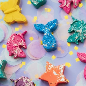 May include: A collection of shaped wax crayons including a yellow butterfly, a blue mermaid tail, pink and teal unicorn heads, and an orange star. Several of the crayons feature silver glitter flecks embedded within the wax, creating a textured appearance.