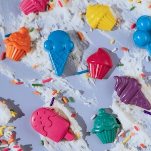 May include: A collection of colourful wax crayons shaped like various desserts, including cupcakes, ice cream cones, and a popsicle. The crayons are scattered across a light grey surface covered in white texture and small, multicoloured sprinkles.